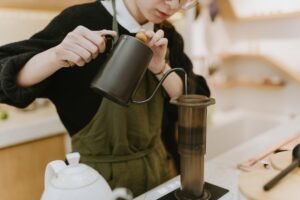 Barista nalewa wodę z czarnego czajnika z wylewką „gooseneck” do zaparzacza typu AeroPress ustawionego na wadze – manualne parzenie kawy w kawiarni.