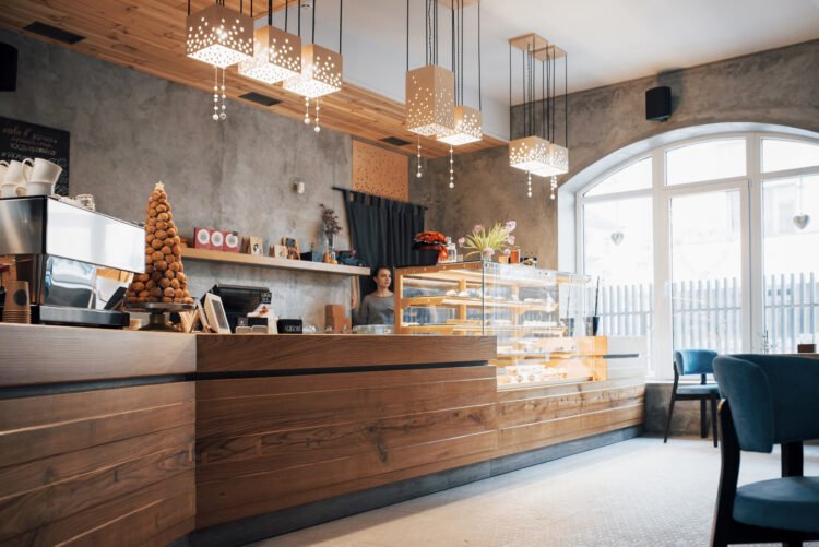 Wnętrze małej kawiarni z długą ladą, ekspresem i przeszkloną witryną z wypiekami; w tle pracownik