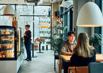 Wnętrze małej kawiarni z ladą ekspozycyjną i gośćmi przy stoliku; w tle obsługa przy barze