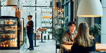 Wnętrze małej kawiarni z ladą ekspozycyjną i gośćmi przy stoliku; w tle obsługa przy barze