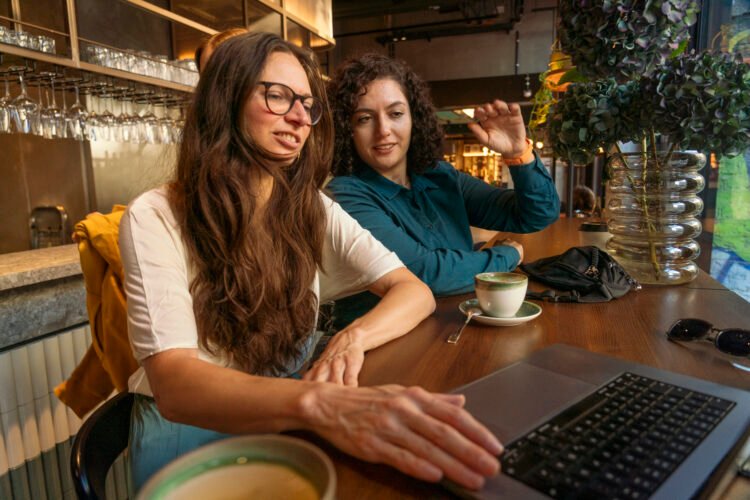 Dwie kobiety przeglądają coś na laptopie w kawiarni; na stole stoją filiżanki kawy i widać bar w tle