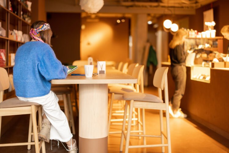 Klientka pracuje przy stoliku w kawiarni z tabletem i kawą; w tle lada z obsługą i witryną